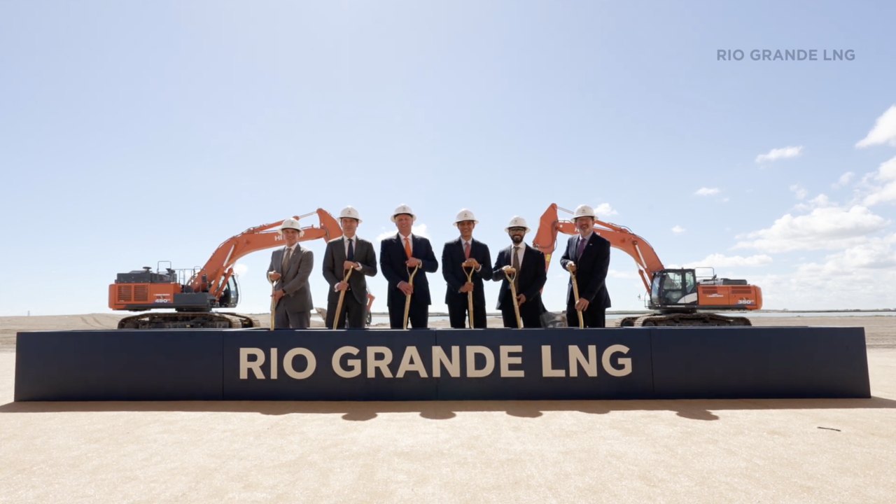 Six executives in hard hats turning ceremonial shovels in front of the branded wall and excavators