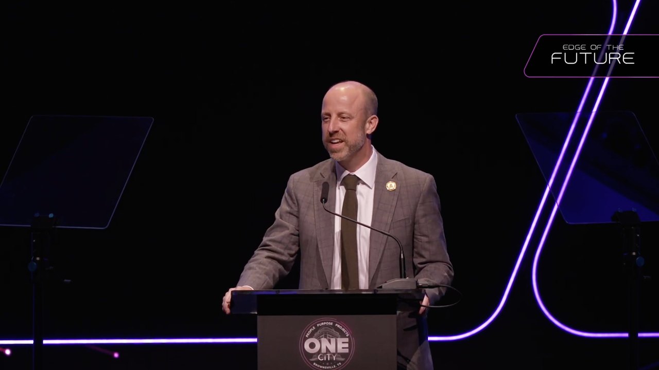 A speaker at the podium with the event's branded mark in the upper corner of the broadcast