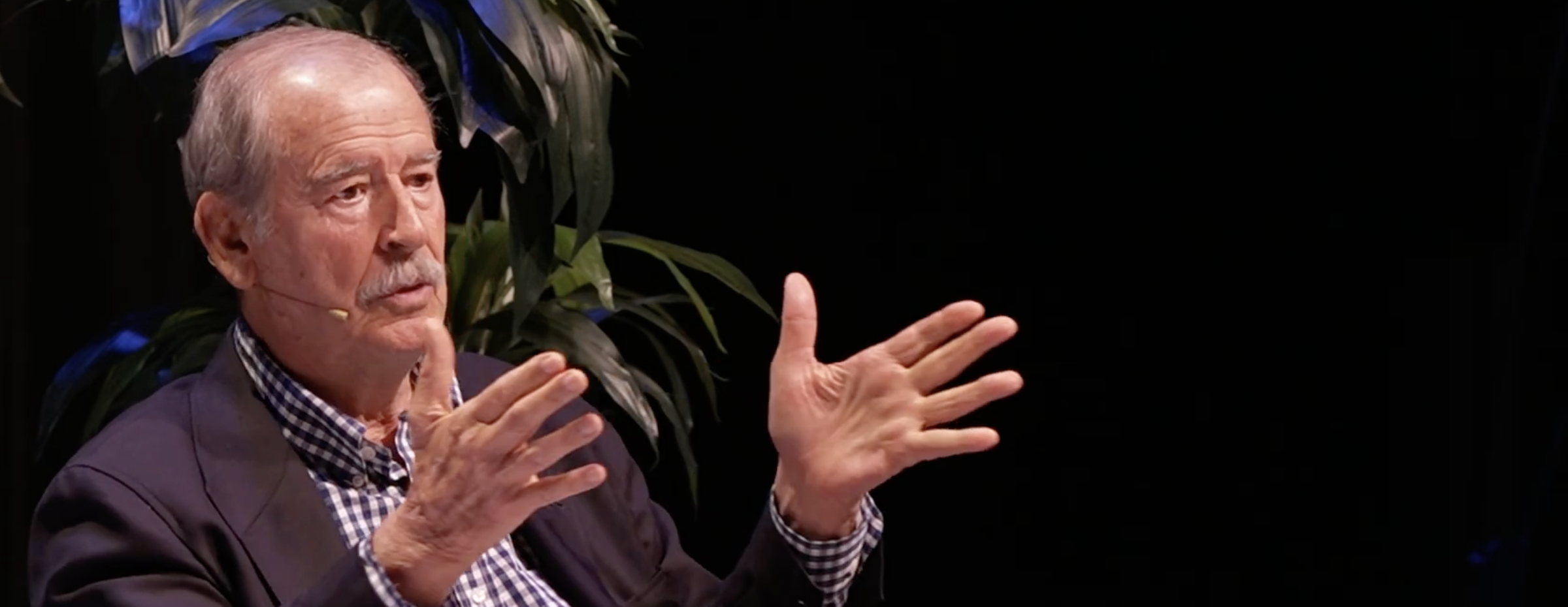 A keynote guest speaking in a seated onstage conversation, lit against a dark theater background