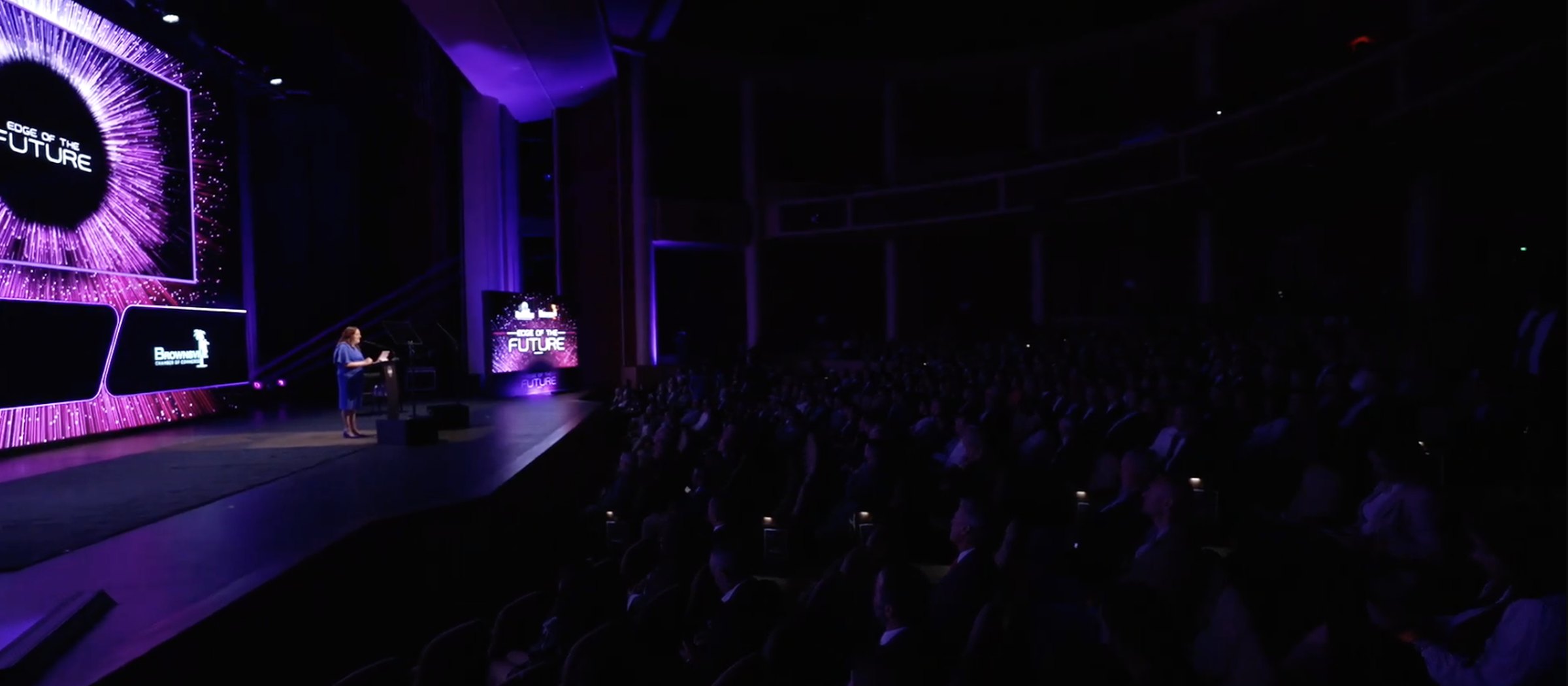 A full house watching a keynote on a purple-lit stage with the show's custom graphics on the main screen