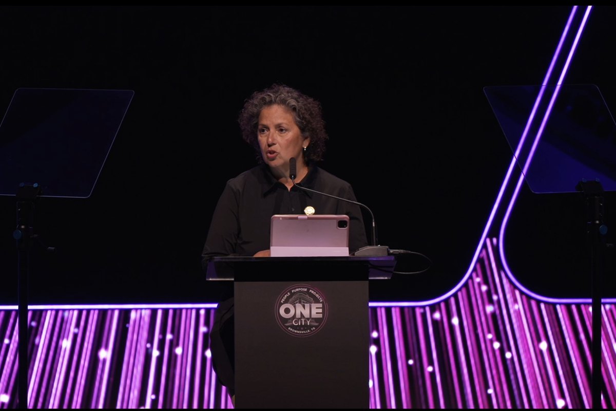 A speaker at a lit civic podium with a purple pinstripe LED backdrop and glass teleprompters