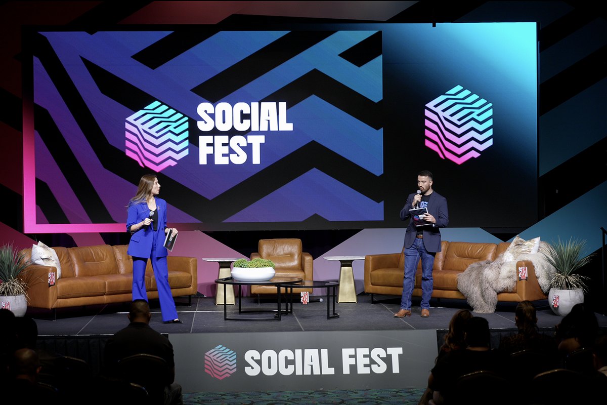 Two hosts on a festival stage in front of a wide LED wall carrying the event's gradient chevron artwork
